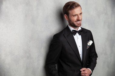 Pozitif genç adam yan bakıyor ve siyah bir smokin giyen ve gri stüdyo arka plan eğilerek ceketinin elini tutarak