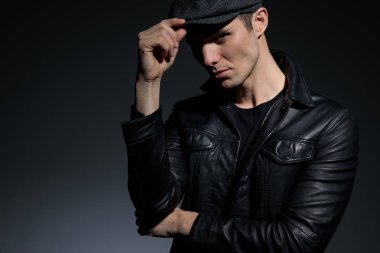 Siyah deri ceket giyip beyaz stüdyo arka planında dururken şapkasını ayarlayan ve kameraya güvenle bakan ciddi görünümlü bir adamı kapatın