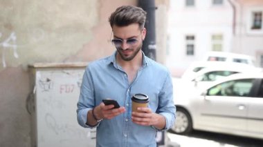 güneş gözlüğü takan genç rahat adam ayakta ve eski bir şehirde, kahve içerken ve gülerken telefonuyla mesajlaşma