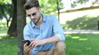 kot gömlek ve güneş gözlüğü genç adam telefonunda mesajları okur ve otururken parkta birini selamlıyor