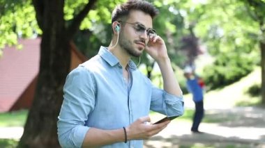 Güneş gözlüğü takan genç mutlu rahat adam parkta yürüyor ve onun telefon müzik dinler , o zevk olarak başını vurma