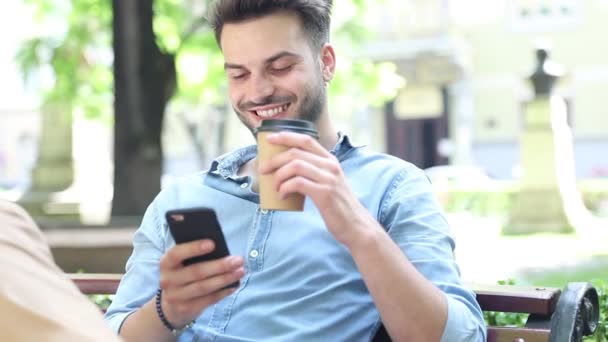 Uvolněný příležitostný muž, který odpočívá na lavičce v parku a používá svůj elegantní telefon, usměje se a pije kávu — Stock video