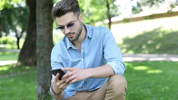 Young handsome man texting on his phone and waves hello at someone in the park — Stock Video