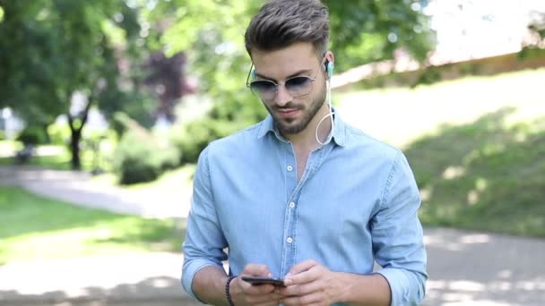 Giovane uomo bello casual con gli occhiali da sole sta camminando nel parco e ascolta la musica sul suo telefono e si guarda intorno — Video Stock