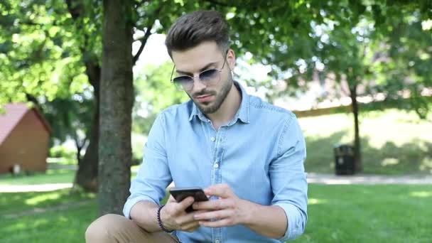 Young casual man sits in the park and writes messaes on his phone — Stock Video