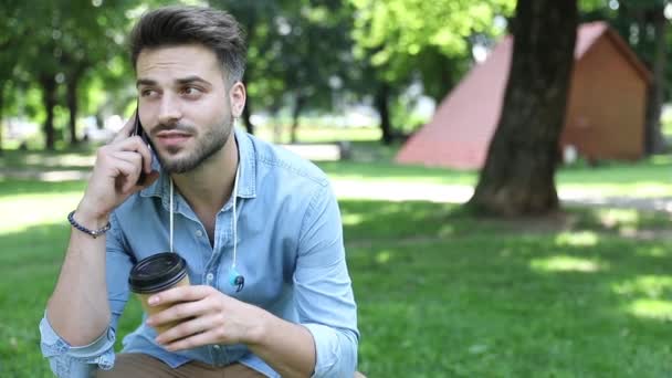 Šťastný muž sedí a mluví po telefonu, zatímco si pije ranní kávu v parku — Stock video