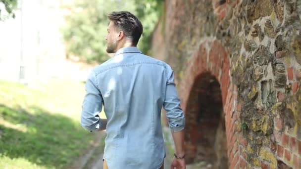 Vista trasera de un hombre casual caminando fuera cerca de la vieja pared de ladrillo, lo llaman y se gira para saludar y saludar a alguien — Vídeos de Stock