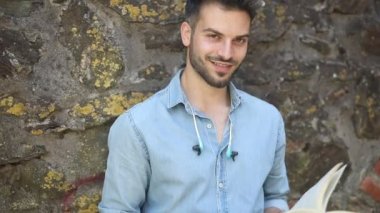 mutlu gülümseyen rahat adam bir kitap okumak ve kamera göz kırpıyor , açık havada eski vintage tuğla duvara karşı ayakta