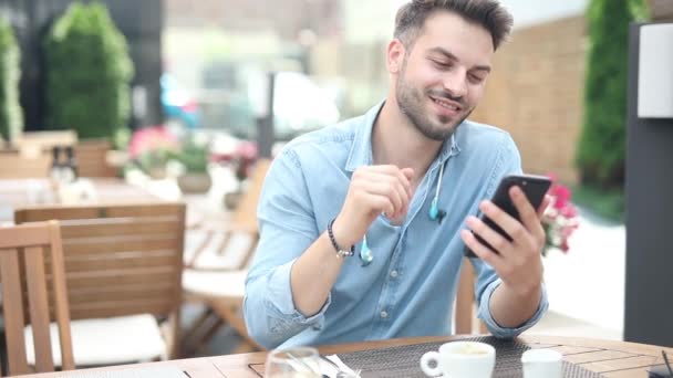Uomo casuale sorridente si siede al ristorante e legge nutrirsi del suo telefono cellulare mentre beve il suo caffè e si gode il suo tempo libero — Video Stock