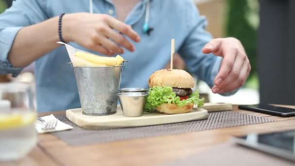 Περιστασιακή άνθρωπος δεν μπορεί να αποφασίσει πώς να αρπάξει και να φάει το μεγάλο χάμπουργκερ του, ενώ κάθεται σε ένα τραπέζι εστιατορίου, υπαίθριο — Αρχείο Βίντεο