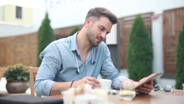 Uomo casuale sorridente si siede al ristorante e legge nutrirsi sul suo tablet mentre beve il suo caffè e si gode il suo tempo libero — Video Stock