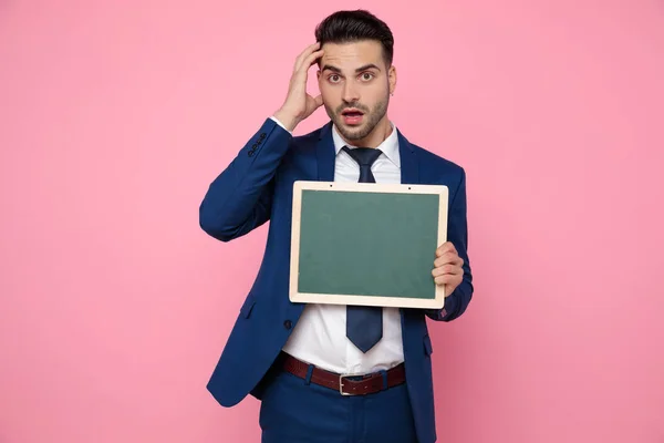 Confused young man holding an empty board on pink background — Stock Photo, Image