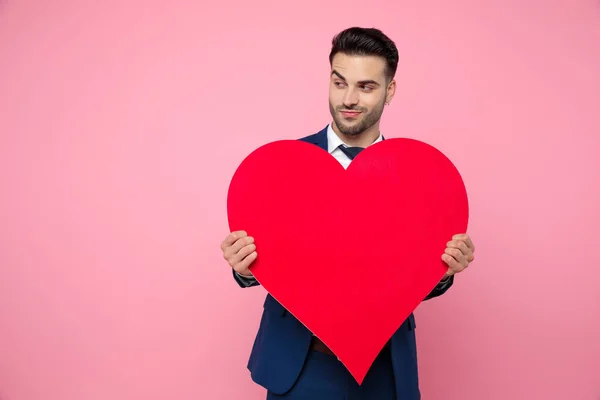 Attraente giovane uomo che tiene il cuore rosso e in piedi sulla schiena rosa — Foto Stock