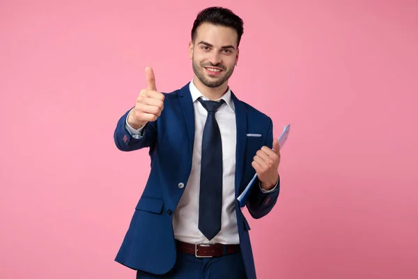 Schöner junger Mann hält Klemmbrett und macht Daumen hoch Zeichen — Stockfoto