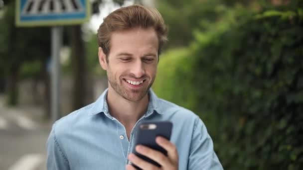 快乐英俊的金发男子走和阅读消息在他的手机上 他微笑着 环顾四周 户外在人行道上 — 图库视频影像