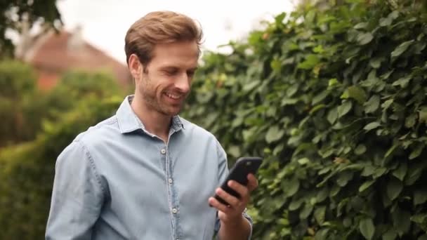 微笑的休闲男子在街上走 并发短信在他的手机 — 图库视频影像