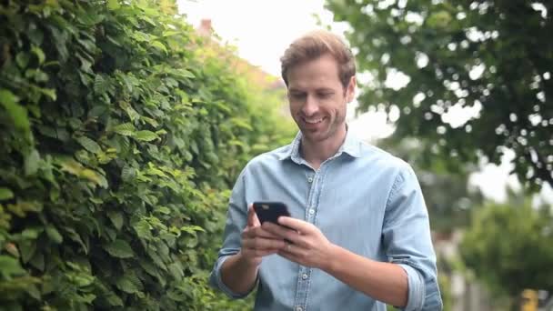 Uśmiechnięty Młody Casual Man Czytania Wiadomości Jego Telefon Fale Rękę — Wideo stockowe