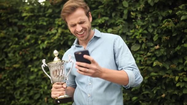 笑的年轻人读好消息在他的手机 而拿着一个大奖杯杯 — 图库视频影像