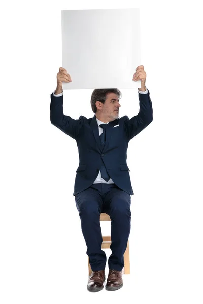Бизнесмен сидит и держит билборд накладные расходы во время просмотра — стоковое фото