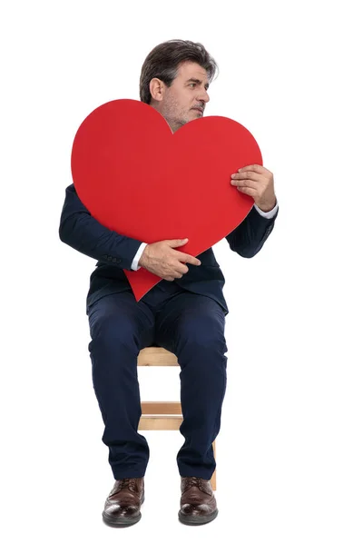 Homem de negócios sentado e segurando um grande coração vermelho olhando para os lados — Fotografia de Stock