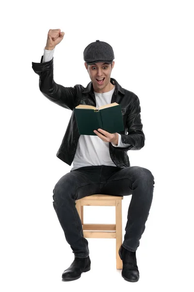 Sentado hombre casual leyendo un libro con el puño en alto emocionado — Foto de Stock