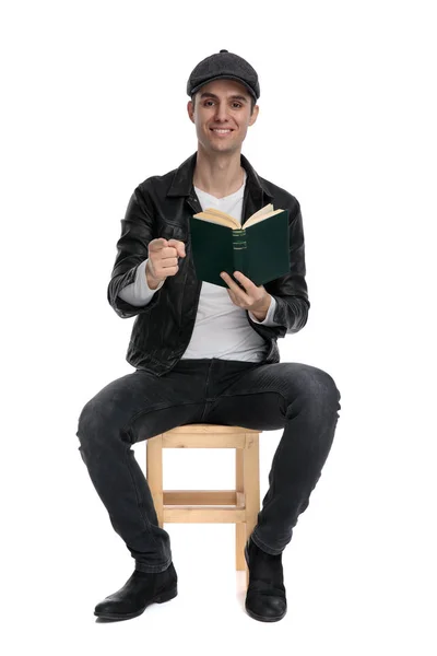Sentado hombre casual sosteniendo libro mientras señala hacia adelante confiado —  Fotos de Stock