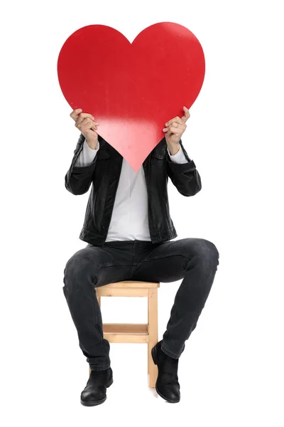 Sitzender lässiger Mann, der sein Gesicht mit einem großen roten Herz bedeckt — Stockfoto