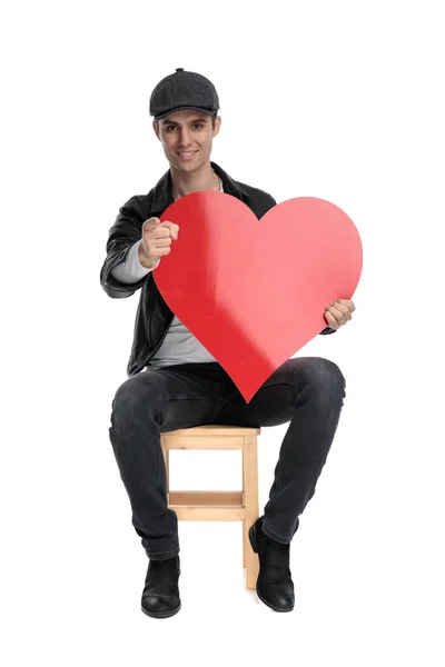 Sentado hombre casual sosteniendo un gran corazón rojo apuntando hacia adelante — Foto de Stock