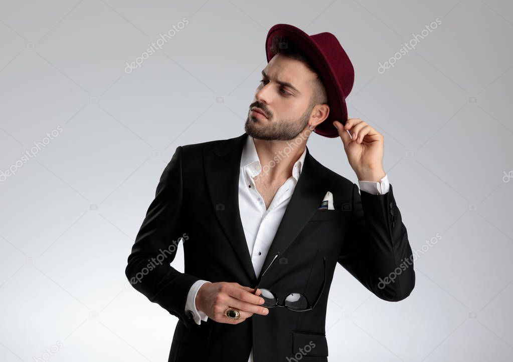 businessman holding glasses and fixing hat while looking away pe