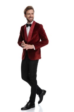 Gülümseyen damat kırmızı kadife smokin giyiyor ve el ele tutuşuyor.