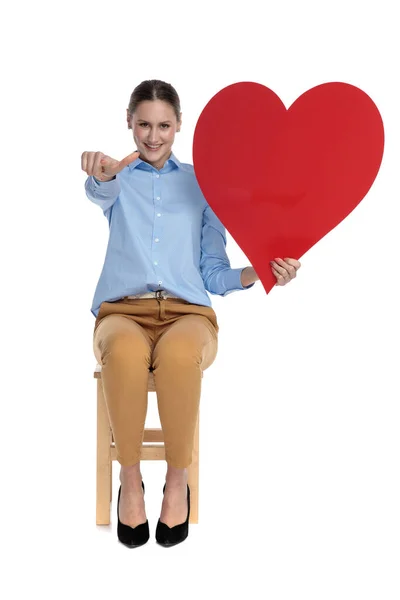 Genç bir kadın işaret ediyor ve büyük kırmızı bir kalp tutuyor. — Stok fotoğraf