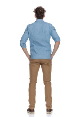 Arka planda, cebinde el ele tutuşan, beyaz arka planda tek başına duran, tüm vücudunu saran genç bir adam var.