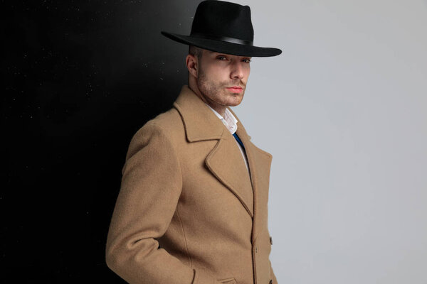 вид сбоку уверенного в себе молодого парня в длинном пальто, позирующего и стоящего на черном сером фоне
