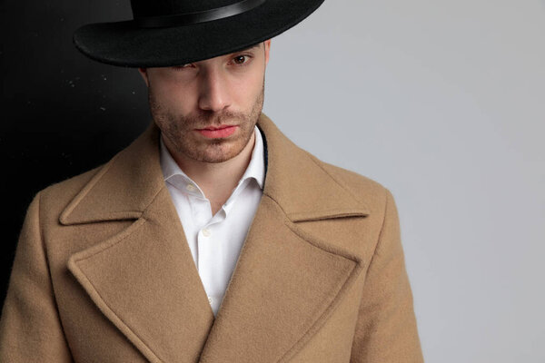 крупным планом молодой модели в длинном пальто в шляпе, позирующей и стоящей на черно-сером фоне
