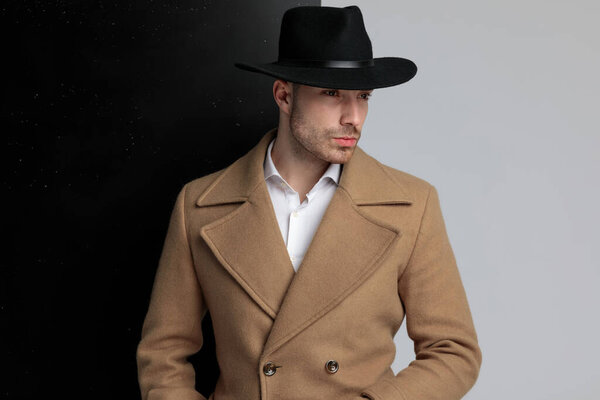 молодой элегантный парень в длинном пальто смотрит в сторону и позирует, стоя на черном сером фоне
