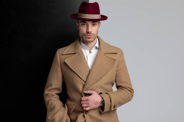 сексуальный молодой человек в шляпе и регулируя длинное пальто, стоя на черном сером фоне
