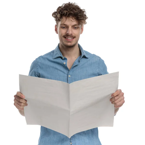 デニムシャツを着た幸せな若い男が新聞を読んで白い背景に孤立して立って肖像画 — ストック写真