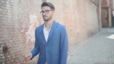 Genç, akıllı, sıradan iş adamı kolunu düzeltiyor, yürüyor, bir elini cebine sokuyor, gözlüklerini tamir ediyor ve el sallıyor.