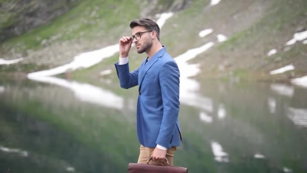 Inteligentny Dorywczy Przystojny Biznesmen Trzymający Walizkę Naprawiający Okulary Spacerujący Jeziorze — Wideo stockowe