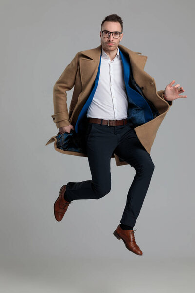 smartcasual young man wearing long coat and jumping in the air on grey background