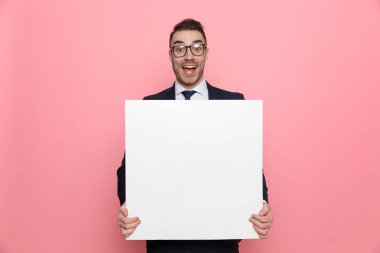 Takım elbiseli, gözlüklü, boş tahta tutan, gülümseyen, pembe arka planda duran mutlu bir iş adamı.