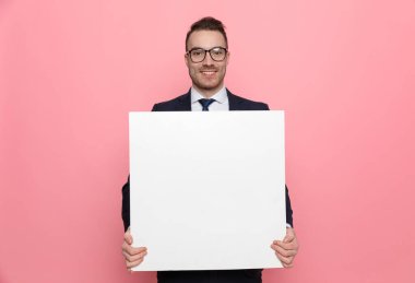 Mutlu genç iş adamı beyaz boş tahtayı sunuyor ve gülümsüyor, pembe arka planda duruyor.