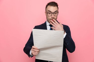 Şok olmuş takım elbiseli, gözlüklü, ağzı eliyle kaplayan, gazete okuyan, pembe arka planda duran şık bir adam.