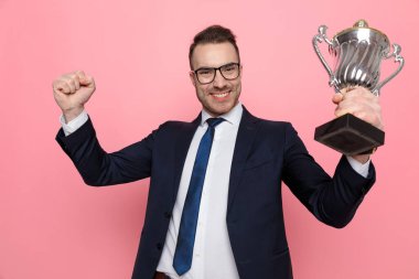 Gözlük takan, havada yumruk ve kupa tutan, zaferi kutlayan, gülümseyen ve pembe arka planda duran mutlu hevesli adam.