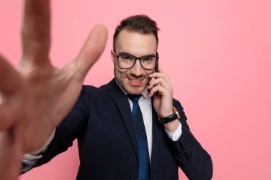 Takım elbiseli mutlu genç işadamı gözlük takıyor, telefonla konuşuyor ve gülümsüyor, pembe arka planda duruyor.