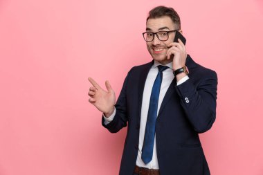 Takım elbiseli, gözlüklü mutlu bir genç. Parmakla işaret ediyor ve telefonla konuşuyor. Pembe arka planda duruyor.