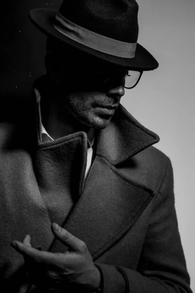 長いコートに眼鏡と帽子をかけファッションのポーズで手を取り黒い灰色の背景の影の中に立つ劇的なエレガントな男 — ストック写真