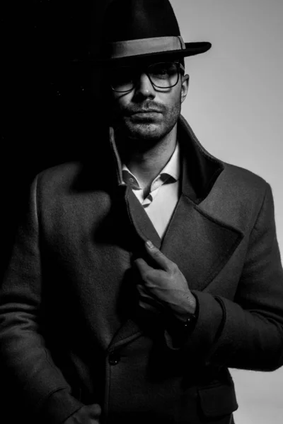 眼鏡と帽子をかけてコートを調整し黒灰色の背景に立つ劇的なクールな男 — ストック写真