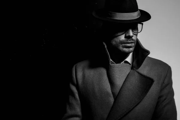 長いコートを着た魅力的なエレガントな男が帽子と眼鏡をかぶって黒い灰色の背景に影の中に立って — ストック写真