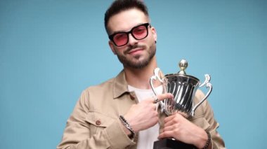 Gururlu, genç, sıradan bir adam kendini ve kamerayı kupa ile gösteriyor. Yumruğunu havada tutuyor ve zaferi kutluyor. Kupa sunuyor ve mavi arka planda hava atıyor.
